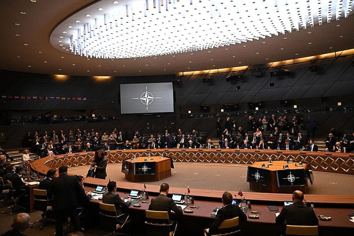 Apvalus posėdžių salės stalas su delegacijomis, ekrane – NATO emblema, aplink sėdi atstovai