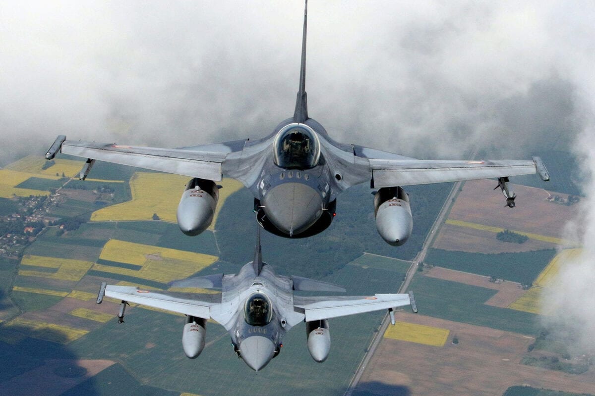 Du naikintuvai F-16 skrenda rikiuotėje virš laukų ir debesų, matomi iš priekio