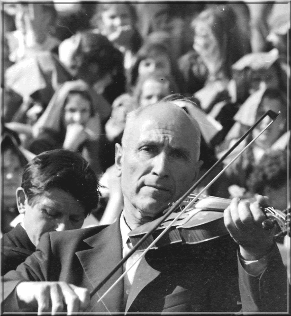 Nespalvota nuotrauka: vyresnis vyras groja smuiku, už jo minia žmonių ir kitas muzikantas