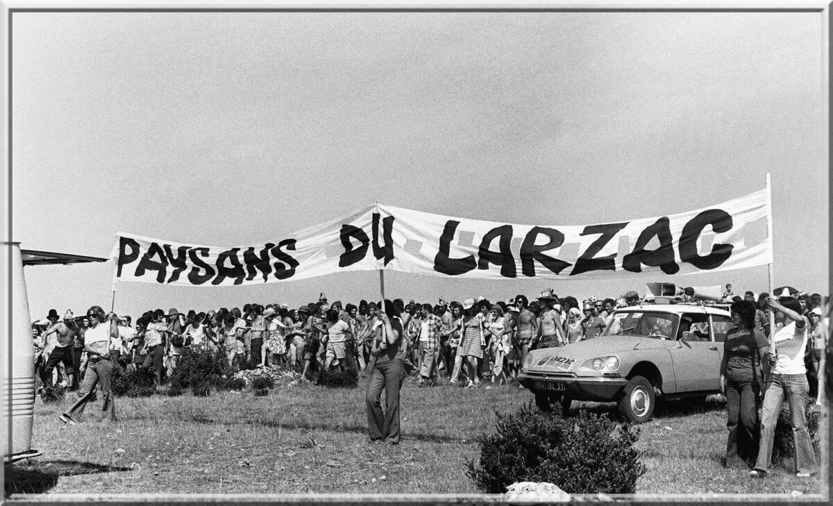 Nespalvota nuotrauka: minia su dideliu plakatu „Paysans du Larzac“ pievoje, šalia senas automobilis
