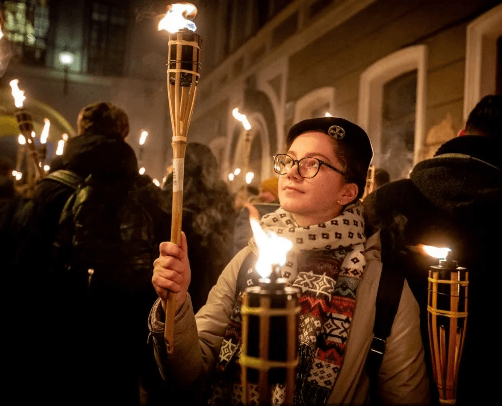 Jauna moteris su berete ir akiniais laiko degantį fakelą naktinėje eisenoje tarp žmonių