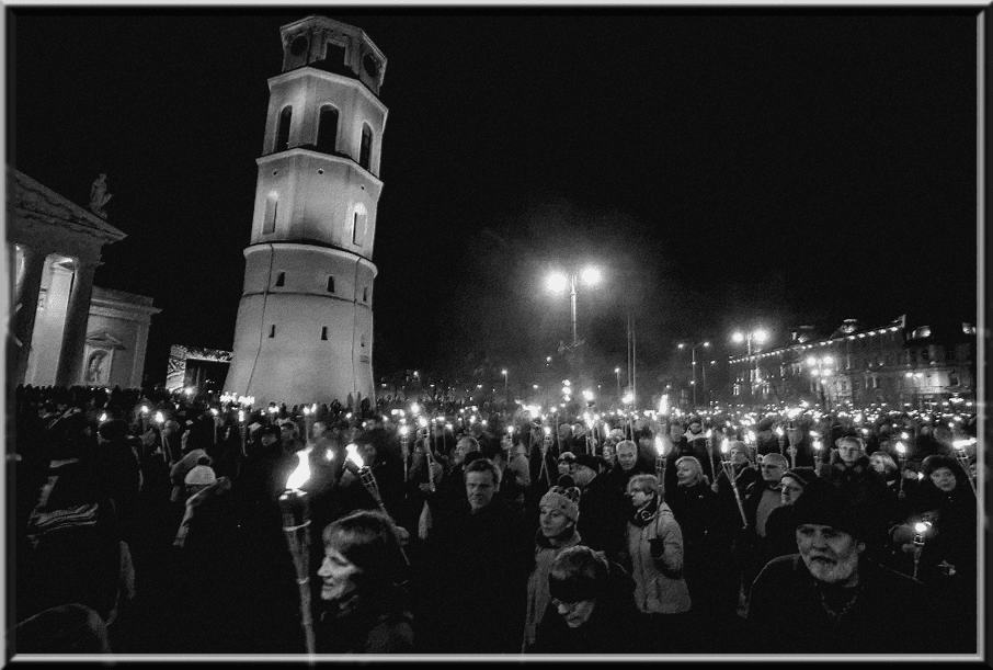 Nespalvota nuotrauka: minia žmonių su deglais naktį prie varpinės bokšto aikštėje